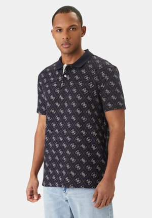 Mann trägt ein schwarzes Polohemd mit weißem Logomuster und hellblauen Jeans, steht vor einem schlichten hellen Hintergrund.