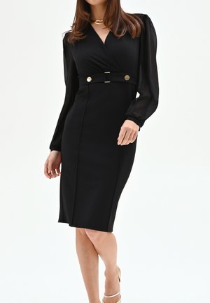 Femme portant une robe noire mi-genou avec des manches longues transparentes, un col en V, une ceinture ajustée avec des boutons dorés, et des talons hauts couleur nude.