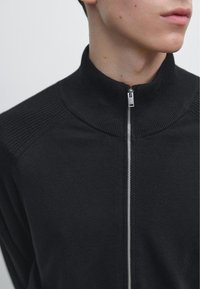 Zwarte trui met rits en hoge kraag. Heeft een geribbelde textuur bij de kraag en schouders, gladde stof over het geheel en zilveren ritsdetail.