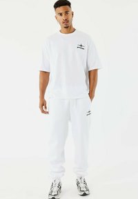 T-shirt bianca in cotone e pantaloni jogger abbinati presentano un logo nero sul petto e sul lato. I pantaloni hanno una vestibilità rilassata e polsini. Sneakers nere e bianche.