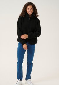 Svart fleecejacka med hög krage, dragkedjestängning och texturerad yta. Bärs över blå jeans och vita sneakers, med händerna vilande på höfterna.