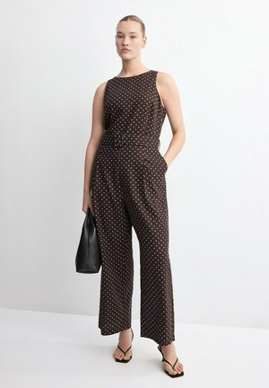 Mujer con mono marrón sin mangas con lunares y cinturón, sosteniendo un bolso negro, de pie contra un fondo blanco liso.