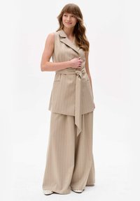 Beige gestreepte, mouwloze utility vest met een strikceintuur, gecombineerd met losse wijde broekspijpen, beide in een gestructureerde stof.