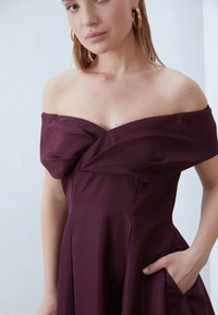 Μπορντό φόρεμα με ανοιχτούς ώμους, με πτυχωτό γιακά, πτυχωτή φούστα και πλαϊνή τσέπη. Λείο ύφασμα με απαλή υφή. Ορατά χρυσά σκουλαρίκια κρίκοι.