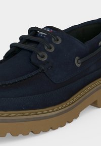 Σκούρο μπλε δερμάτινο παπούτσι τύπου boat shoe με παχιά ταμπά λαστιχένια σόλα, μαύρα κορδόνια διά μέσου μεταλλικών οπών και ορατή ετικέτα με το λογότυπο στη γλώσσα.