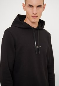 Black hoodie made of soft fabric, featuring a drawstring hood, ribbed cuffs, and a small logo on the chest. Smooth texture throughout.