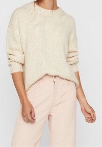 En kvinna bär en mjuk, löst sittande krämfärgad tröja och ljusrosa kordbyxor, med händerna vilande på höfterna.