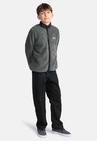 Grauer Fleece-Zip-Jacke mit schwarzem Kragen, getragen über leicht sitzenden schwarzen Hosen und grauen Sneakers. Mit einem kleinen Logo auf der Brust.