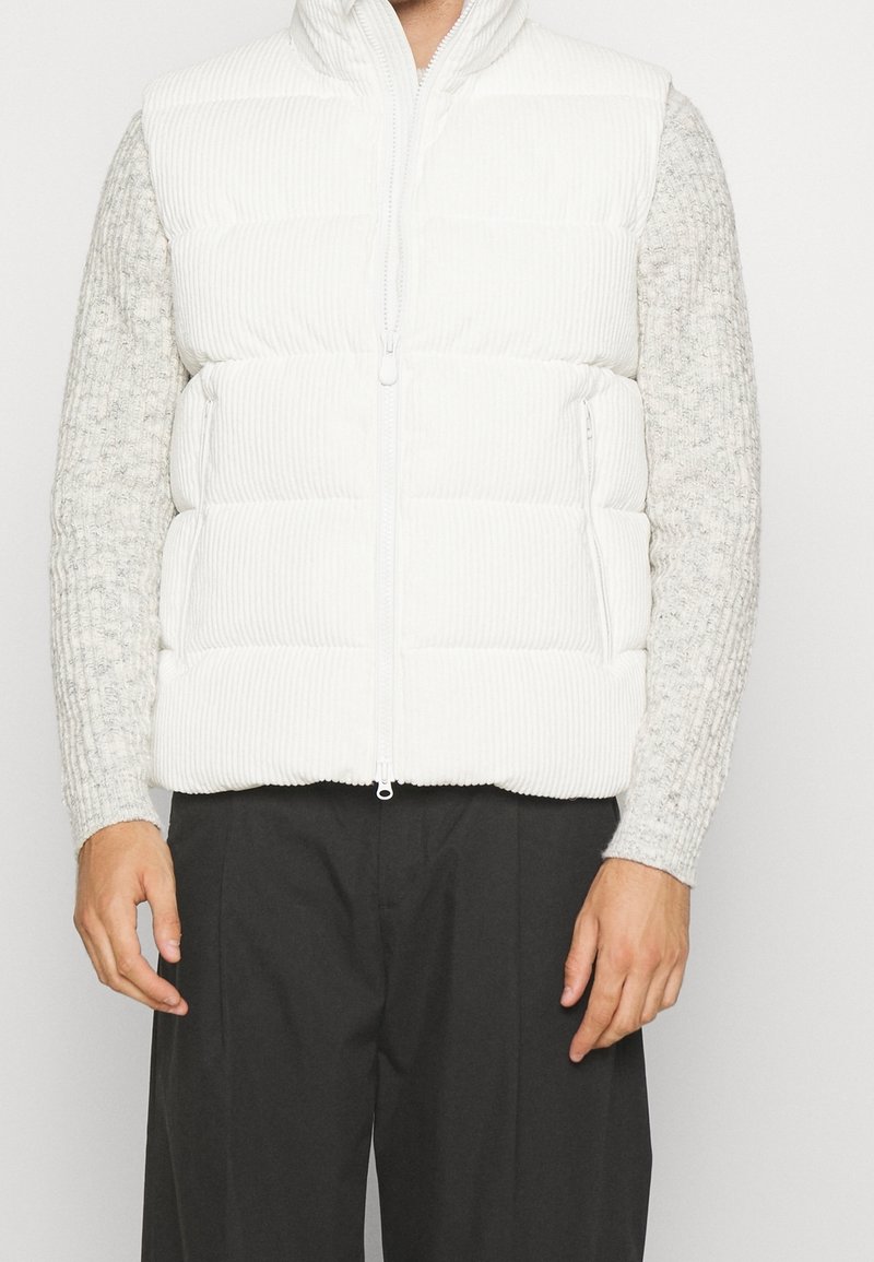 Homme portant une doudoune en velours côtelé blanc sur un pull en tricot gris clair et un pantalon noir, debout devant un fond uni.