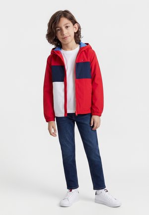 Dziecko z brązowymi włosami do ramion, ubrane w czerwoną, białą i granatową kurtkę z kapturem, białą koszulkę, ciemne dżinsy oraz białe trampki.