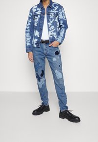 Jeansjacka med graffiti-mönster, ljusblå jeans med lappar i olika nyanser av blått, parat med svarta snörskor.