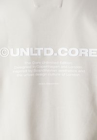 Lys grå sweatshirt med stort hvidt teksttryk. Teksten inkluderer "©UNLTD.CORE" og en beskrivelse af sin designinspiration.