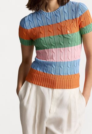 Vrouw draagt een kortarmige kabelgebreide trui met horizontale strepen in blauw, oranje, groen en roze, gecombineerd met witte hoge taillebroeken.