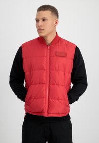 Veste rembourrée rouge avec fermeture éclair sur le devant, design matelassé et col côtelé. Présente une étiquette marron sur la poitrine et est portée par-dessus une chemise à manches longues noire.