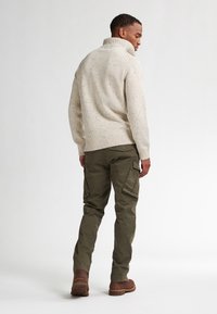 Maglione a maglia color crema con colletto alto, texture screziata; abbinato a pantaloni cargo verde oliva e stivali marroni, visto dal retro.
