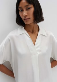 Blusa bianca a maniche corte con colletto, caratterizzata da una scollatura a V e un tessuto liscio e leggero. Il design è semplice ed elegante.