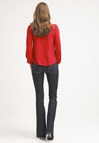 Röd blus med lång ärm och åtsittande manschetter, kombinerad med mörk indigoblå utsvängda jeans. Mjukt tyg och subtila sömnadsdetaljer.