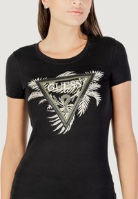 T-shirt nera a maniche corte con una grafica triangolare con la scritta "GUESS", motivi di foglie e accenti argentati. Realizzata in morbido cotone.