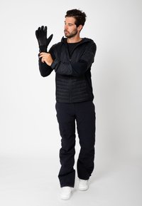 Veste isolante noire avec fermeture éclair à l'avant, portée avec des gants noirs et un pantalon noir. Le modèle ajuste le gant de sa main gauche.