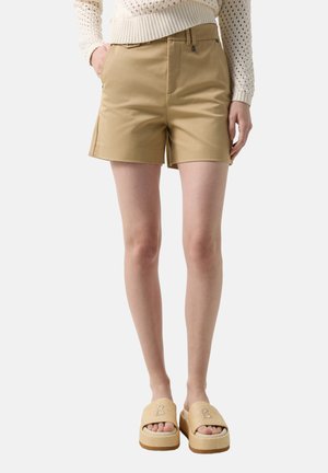 Kobieta w beżowych szortach z wysokim stanem, kremowym swetrze dzianinowym i beżowych klapkach na platformie, stojąca z częściowo wsuniętymi rękami do kieszeni.