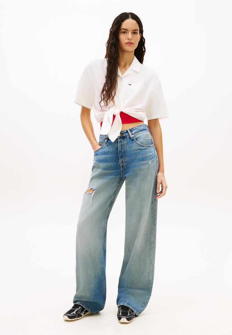 Γυναίκα με μακριά κυματιστά μαλλιά που φοράει λευκό πουκάμισο με κοντά μανίκια δεμένο μπροστά πάνω από κόκκινο τοπ, φαρδιά τζιν παντελόνια με σκισίματα και μαύρα αθλητικά παπούτσια.