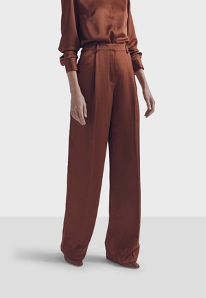 Persona che indossa pantaloni marroni larghi a vita alta e un top a maniche lunghe abbinato, in piedi davanti a uno sfondo chiaro e uniforme.