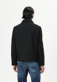 Black unstructured coat with a notched collar, shown from the back. Worn with blue denim jeans featuring angled back pockets.