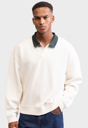 White pullover sweatshirt with a dark green collar, relaxed fit, ribbed cuffs and hem, worn over blue denim jeans. Smooth texture, no patterns.