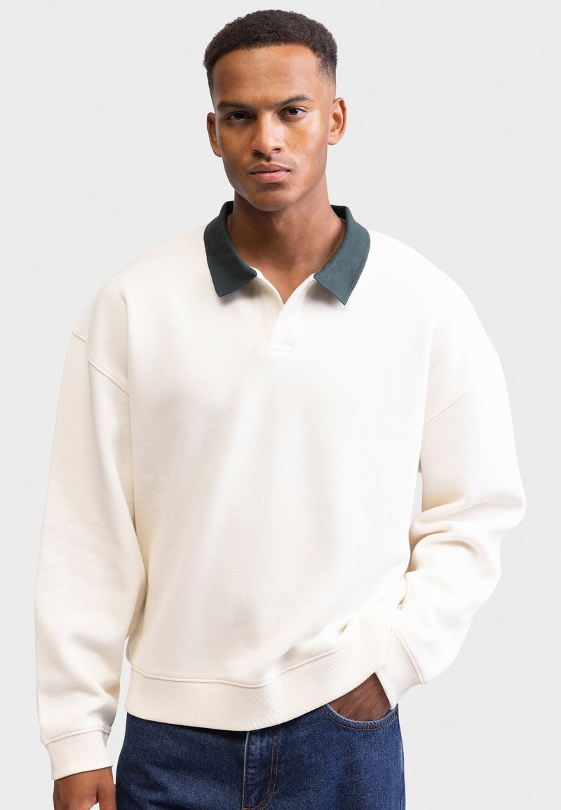 White pullover sweatshirt with a dark green collar, relaxed fit, ribbed cuffs and hem, worn over blue denim jeans. Smooth texture, no patterns.