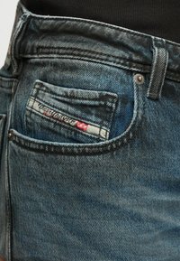 Plano de perto de um bolso de jeans de denim azul com etiqueta Diesel Industry e detalhes de costura visíveis.