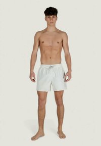 Jaunas vyras stovi basas, be marškinėlių, vilkintis baltus maudymosi šortus su šonu kišene prieš vienodą foną.