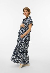 Femme enceinte portant une robe à motifs bleus et blancs, tenant son ventre, debout devant un fond blanc uni.