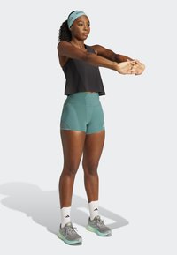 Sort cropped tank top, teal højtaljede shorts, grå løbesko med mint-accenter og hvide sokker med sorte striber. Strækningsposition.