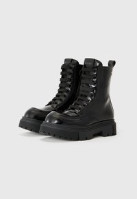 Bottines en cuir verni noir avec une semelle épaisse, un laçage haut et une tirette à l'arrière. Présente des lacets noirs texturés et un détail signature.