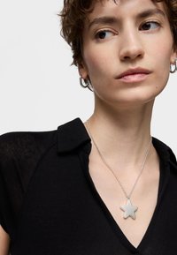 Collana con pendente a forma di stella argentata su una catena sottile, indossata sopra una maglietta nera con colletto. La stella è liscia e ha una finitura opaca.
