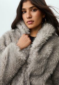 Giovane donna con lunghi capelli scuri che indossa grandi orecchini a cerchio e un cappotto di pelliccia sintetica grigia chiara e soffice, mentre tiene il collo del cappotto con una mano.