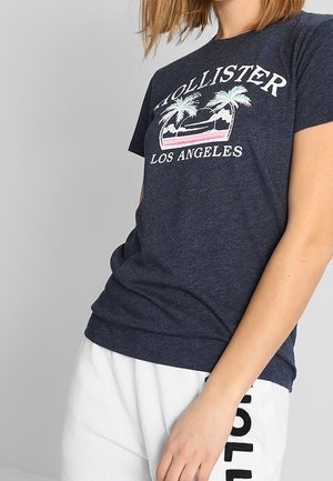 Person wearing a navy Hollister Los Angeles t-shirt with palm tree graphic and white shorts with black text on side.