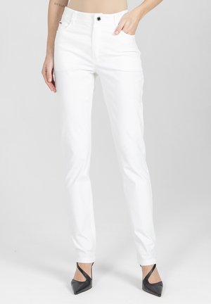 Kobieta w białych jeansach o prostych nogawkach i czarnych czółenkach na wysokim obcasie ze spiczastym noskiem, stojąca na tle jednolitego jasnoszarego tła.