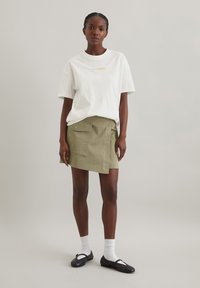 Hvid bomulds t-shirt med orange tekst, parret med en grøn, knælang omslagsskjørt med en sidelomme og knapdetalje. Sorte sko og hvide sokker.