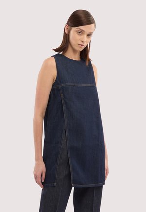 Kobieta o prostych brązowych włosach nosi bezrękawnik z ciemnoniebieskiego denimu z rozcięciami po bokach oraz pasujące ciemne dżinsowe spodnie na jasnym tle.