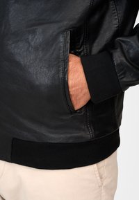 Black leather jacket with a smooth texture, zippered pockets, and ribbed black cuffs. Featured on a neutral background.