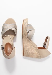 Beige wedge-sandalen met een gegolfde espadrille-zool, voorzien van kruislings lopende banden in metallic zilver met branding. Verstelbare enkelgesp.