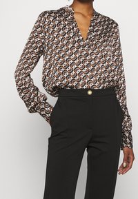 Brun och vit mönstrad långärmad blus med V-hals och något lös passform, parat med högmidjade svarta byxor med en guldbutton.