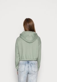 Sudadera corta verde con capucha, hecha de material suave, con hombros caídos y cintura elástica, combinada con jeans de un azul claro.