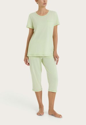 Donna che indossa una maglietta a maniche corte verde chiaro con taschino sul petto e pantaloni corti coordinati, in piedi a piedi nudi su uno sfondo uniforme.