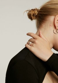 Anillo de plata en una mano tocando un hombro, llevado con una blusa negra. Pendiente con piedras visible en la oreja, cabello recogido en un moño. Textura de piel suave.
