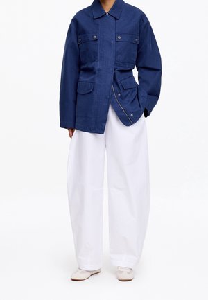 Personne portant une veste bleu marine boutonnée avec des poches à rabat et un pantalon blanc large et ample avec des chaussures blanches sur un fond uni.