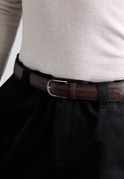 Brun læderbælte med tekstureret overflade, sølvfarvet metalspænde, båret over sorte stofbukser, parret med en lys beige sweater.