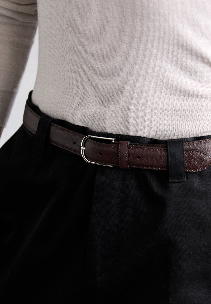 Brown leather belt with a textured finish, silver metal buckle, worn over black fabric pants, paired with a light beige sweater.