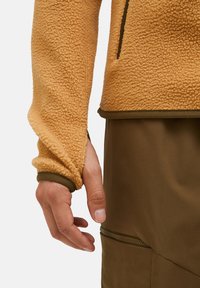 Fleecejacke in Orange mit einer strukturierten, flauschigen Oberfläche; mit braunen Akzenten und einer Reißverschlusstasche an dem braunen unteren Kleidungsstück.